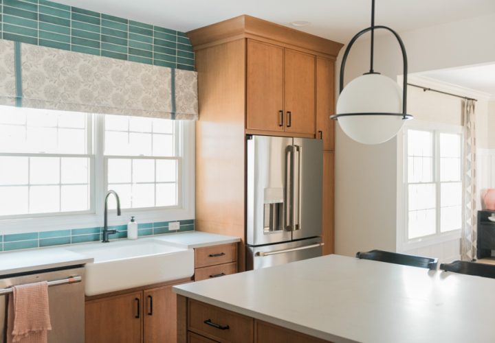 A serene kitchen sink zone with a white apron-front farmhouse sink, matte black gooseneck faucet, and bright window view above — framed by a vibrant teal tile backsplash and warm wood cabinetry for a fresh, inviting feel.